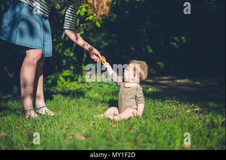 Ein süßes kleines Baby sitzt auf dem Gras außerhalb und nimmt die Hand seiner Mutter Stockfoto