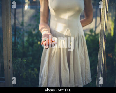 Eine junge Frau trägt ein Kleid steht in Ihrem Gewächshaus bei Sonnenuntergang mit einige Tomaten in Ihrer Hand Stockfoto