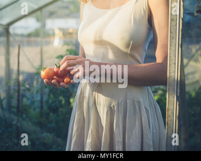 Eine junge Frau trägt ein Kleid steht in Ihrem Gewächshaus bei Sonnenuntergang mit einige Tomaten in Ihrer Hand Stockfoto