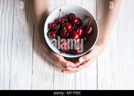 Köstliche reife Kirschen in eine Schüssel in Kinderhand auf weißem Holz- Hintergrund. Stockfoto