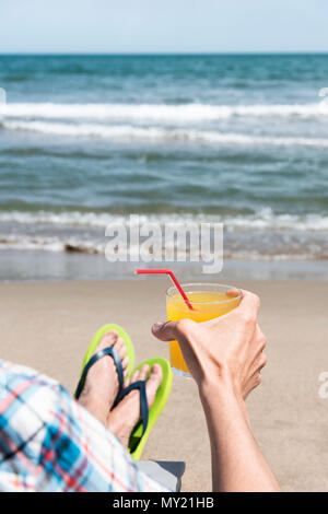 Nahaufnahme eines jungen kaukasischen Mann entspannt am Strand, Sitzen auf einem Liegestuhl und tragen ein Paar der Grünen flip-flops, mit einem Glas mit einem erfrischenden Stockfoto