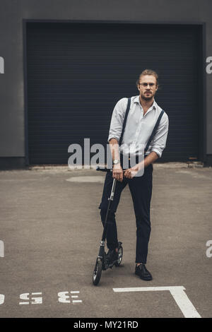 Stilvolle junger Mann in Brillen und Strapsen auf Scooter auf Parkplatz Stockfoto
