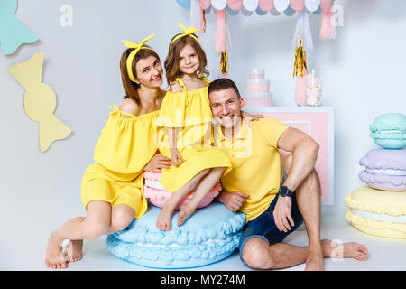 Mama, Papa Umarmungen Tochter, sitzen auf großen makronen vor dem Hintergrund der weißen Wand und Candy Bar im Studio. Gefühle des Glücks. Glückliche Familie po Stockfoto