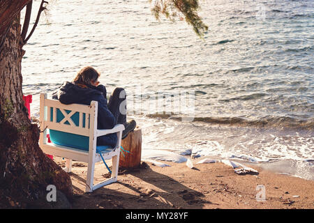 Mittleres Alter kaukasischer Mann sitzt auf einem Stuhl in einer entspannten Position am Meer. Winter auf einem sonnigen Tag. Rückansicht. Stockfoto