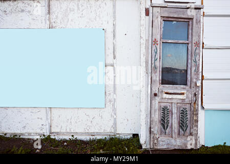 Vintage Holztür eines weißen alten Hütte. Blume Schnitzereien an der Tür und eine große blaue Leere Repräsentant mit Kopie Raum an der Wand hängen. Stockfoto