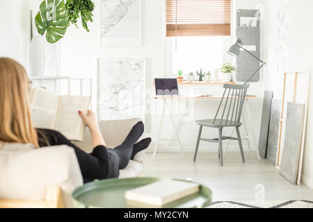 Frau auf dem Sofa und liest ein Buch in Unscharfer Vordergrund in Weiß home office Interieur mit grau Stuhl stehend durch eine hölzerne Schreibtisch mit Lampe und Stockfoto