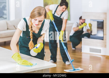 Junge Mitglied einer Reinigung Mannschaft tragen grüne Overalls und gelben Handschuhen wischte einen weißen Tisch in der Wohnung Interieur mit dem Rest der Mannschaft in der Unschärfe Stockfoto