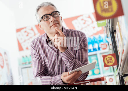 Zuversichtlich Mann tut Lebensmittelgeschäft Einkaufen im Supermarkt, er sucht Produkte und bietet mit Anwendungen auf seine digital-Tablette Stockfoto