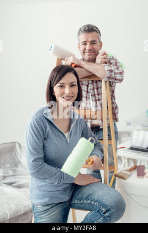 Glückliches Paar renovieren ihr Haus und Malerei ein Zimmer, sie lehnen sich auf einer Leiter und posieren mit Farbe rollen Stockfoto