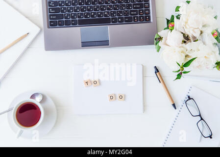 Ansicht von oben Licht arbeiten mit Laptop Tastatur, Notebook zu tun mit Schriftzug, Tasse Tee, sletchbook, Gläser, weiße Pfingstrose auf weißem woodden Tabelle. Arbeitsbereich Konzept. Selektive konzentrieren. Kopieren Sie Platz. Stockfoto