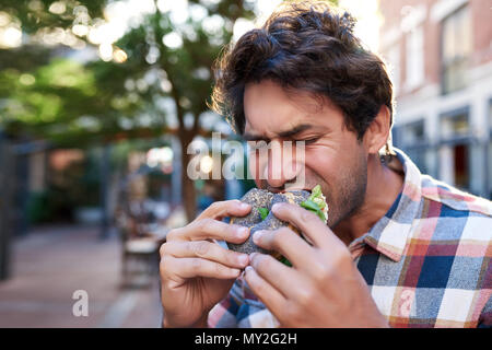 Hungrigen jungen Mann draußen stehen mit geschlossenen Augen und nahm einen Bissen von seinem leckeren Mohn Bagel Stockfoto