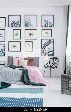 Real Photo von einem breiten Bett steht neben einem schwarzen Stuhl in einem Schlafzimmer Einrichtung mit Pflanzen Poster an der Wand und Teppichen auf dem Boden Stockfoto