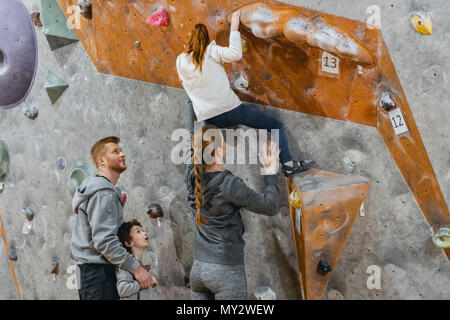 Kleines Mädchen klettern eine Wand mit Grips und ihre Mamma sie Unterstützung von der Rückseite Stockfoto