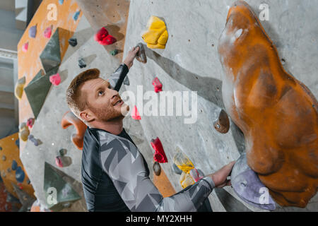 Halbe Länge Schoß des jungen Mannes in sportliche Kleidung klettern eine Wand mit Griffe am Gym Stockfoto