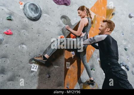 Junge Frau klettern eine Wand mit Grips mit Mann ihr Unterstützung von hinten Stockfoto