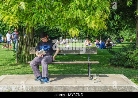 Ein junger erwachsener Mann sitzt auf einer Parkbank mit einem Buch an einem heißen, sonnigen Tag in der Gemeinde Gärten, Hammersmith, London. Stockfoto