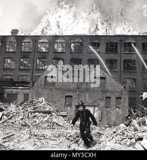 1950er Jahre, historische Bild einer großen Lagerhalle auf Feuer in London, England. Wasser wird auf das brennende Gebäude gesprüht als massive Flammen rip durch das Dach, während ein Feuerwehrmann über die Trümmer, um Unterstützung zu erhalten. Stockfoto