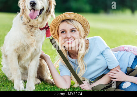 Lächelnde Frau mit Strohhut Golden Retriever Hund streicheln, während auf dem Rasen im Park liegend Stockfoto