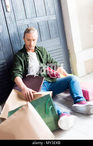 Hübscher junger Mann mit einkaufstaschen beim Stillstehen und sitzen auf dem Boden Stockfoto