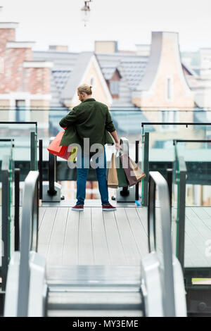 Rückansicht der Mann hält multicolord Papiertüten beim Stehen auf Rolltreppe in Shopping Mall Stockfoto