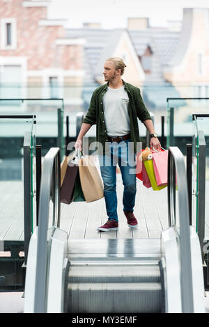 Hübscher junger Mann mit einkaufstaschen beim Stehen auf Rolltreppe und weg schauen Stockfoto