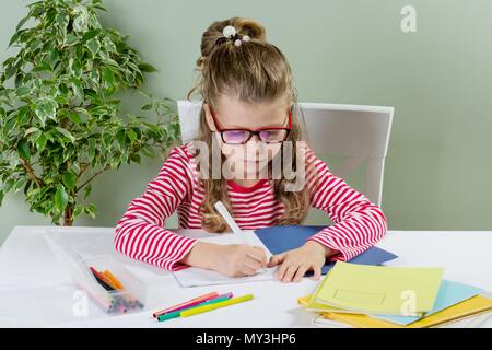 Ein jüngeres Schulmädchen mit Brille schreibt etwas mit der linken Hand auf dem Notebook- und setzt sich an den Tisch. Zurück zum Konzept der Schule. Das Kind ist links Stockfoto