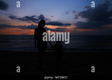 Schwarze Silhouette von Mutter Holding Tochter Hand und Fuß am Meer mit Sonnenuntergang Hintergrund. Stockfoto