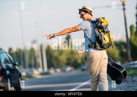 Rückansicht des jungen Mann mit Gitarre Gestik Auto alleine per Anhalter zu stoppen Stockfoto