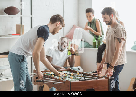 Seitliche Sicht auf zwei junge Männer spielen Tischfußball, während ihre Freunde beobachten mit Bierflaschen Stockfoto