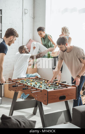 Zwei junge Männer spielen Tischfußball, während ihre Freunde beobachten mit Bierflaschen und Popcorn Stockfoto