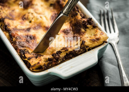 Messer schneidet Lasagne auf dunklen Holztisch Stockfoto
