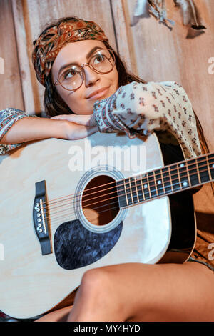 Attraktive hippie Mädchen im Kopfband und Gläsern sitzen mit einer Gitarre in Holz- Haus Stockfoto