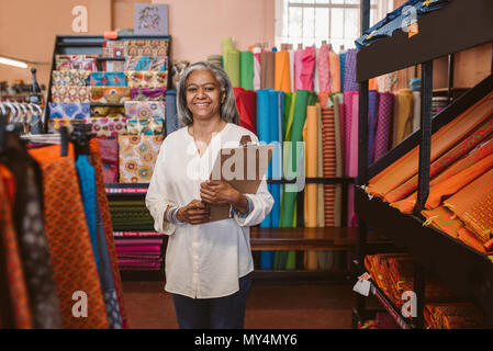 Porträt eines lächelnden reife Stoff shop besitzer eine Zwischenablage beim Stehen neben den Regalen mit bunten Tüchern und Textilien Stockfoto