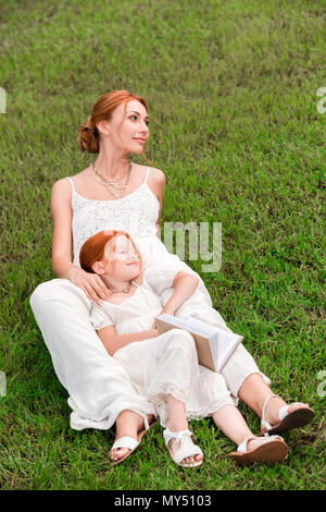 Lächelnd redhead Mutter und Tochter in weißen Kleidern Buch beim Stillstehen auf grünem Gras Stockfoto