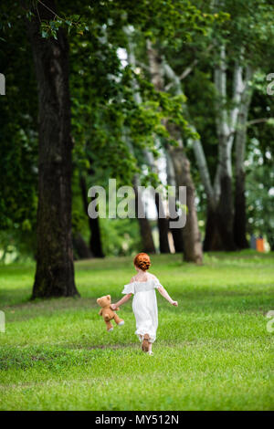 Rückansicht des niedlichen kleinen Mädchen in weißem Kleid holding Teddybären und Wandern auf grünem Gras im Park Stockfoto