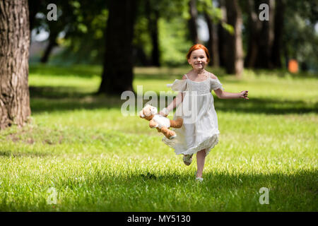 Adorable rothaarigen Mädchen in weißem Kleid holding Teddybären und läuft auf grünem Gras in Park Stockfoto