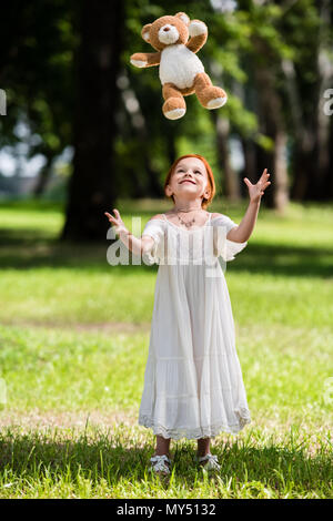 Gerne kleine Mädchen in weißem Kleid werfen Teddybär beim Stehen in Park Stockfoto