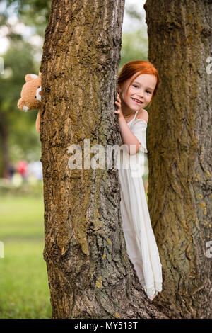 Happy rothaarige Mädchen, dass Teddybären und versteckt sich hinter Baum im Park Stockfoto