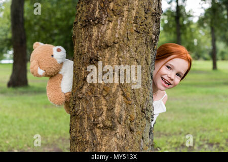 Happy rothaarige Mädchen, dass Teddybären und versteckt sich hinter Baum im Park Stockfoto