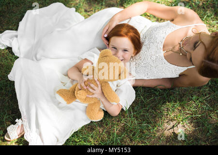 Blick von oben auf die wunderschöne rothaarige Mutter und Tochter mit Teddybär liegend auf grünem Gras in Park Stockfoto