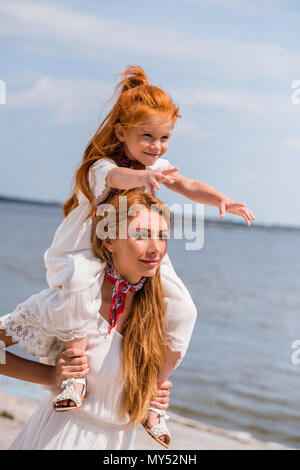 Glückliche Mutter, die entzückende rothaarige Tochter am Hals beim Gehen an der Küste Stockfoto