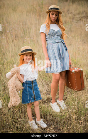 Schönen Mutter und Tochter mit Koffer und Teddybären halten sich an den Händen, während zusammen stehen auf Gras Stockfoto