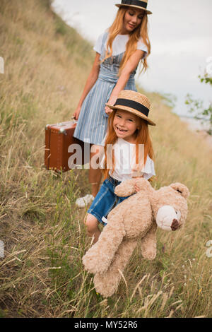 Schöne glückliche Mutter und Tochter holding Koffer und Teddybär beim Gehen auf Grünland Stockfoto