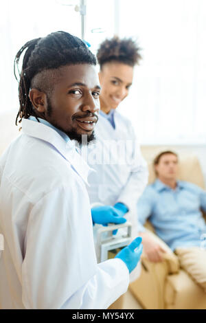 Junge afrikanische-amerikanischen Arzt in Laborkittel und Latex Handschuhe mit seinen Kollegen und Patienten im Hintergrund Stockfoto