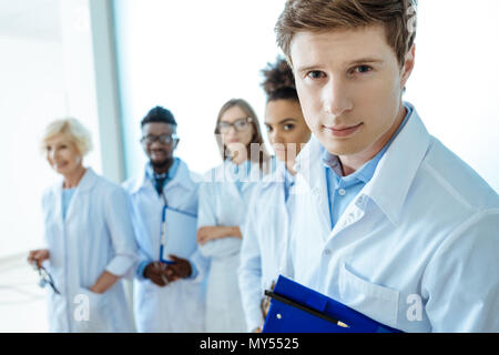 Junger Arzt in Laborkittel holding Zwischenablage mit Gruppe junger Ärzte in Laborkitteln hinter ihm. Stockfoto