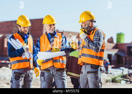 Drei Arbeiter in hardhats Prüfung Baupläne und Sprechen auf tragbaren Radio auf der Baustelle Stockfoto