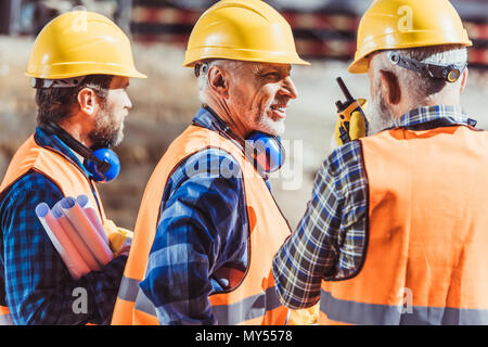 Drei Arbeiter in hardhats und warnwesten an der Baustelle stehen und sprechen über tragbares Radio Stockfoto