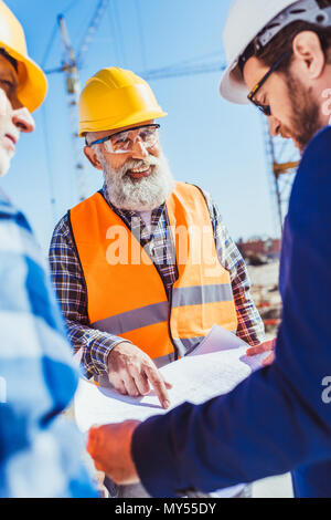 Bauarbeiter in Schützende Uniform diskutieren Baupläne mit Geschäftsmann in hardhat und Anzug Stockfoto