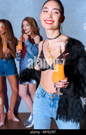 Schönen lächelnden Asiatin holding Glas mit Cocktail, während Freunde hinter Trinken Stockfoto