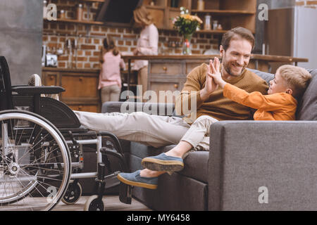 Glücklichen kleinen Sohn mit lächelnden behinderten Vater hoch fünf und Rollstuhl in der Nähe von sofa Stockfoto